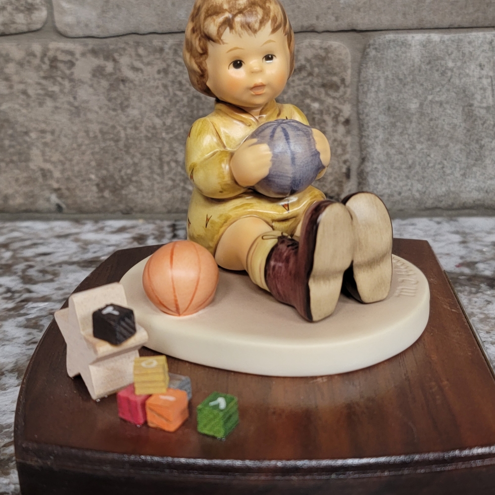 At Play Hummel Figurine with Yellow Outfit and Colorful Toys. Sits On Wood Base